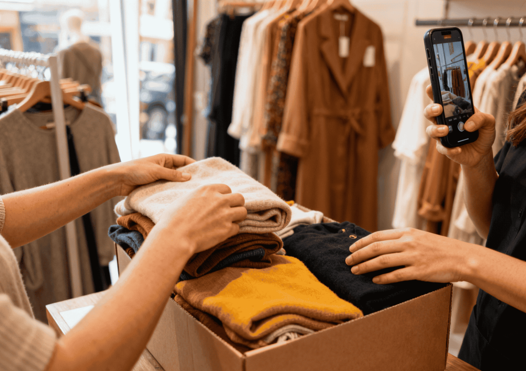 Una imagen hiperrealista y de estilo documental que captura el detrás de cámaras de una pequeña tienda de moda minorista española. Dos mujeres españolas de aspecto natural y concentradas, una con delantal de trabajo, están empacando y preparando pedidos de ropa artesanal en su taller. Ellas están enfocadas en su labor, rodeadas de rollos de tela y herramientas de costura en un entorno de trabajo funcional. A su lado, un teléfono móvil montado en un trípode graba el proceso en directo para un Reels o TikTok, como se muestra en la pantalla del dispositivo. La escena, hiperrealista y de estilo documental, muestra un momento cotidiano de creación de contenido orgánico, transmitiendo humanización de marca, autenticidad y el concepto de marketing de moda Lo-Fi real.