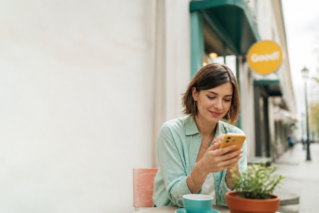 Mujer joven sonriendo mientras consulta su teléfono móvil inteligente en la terraza de una cafetería al aire libre.